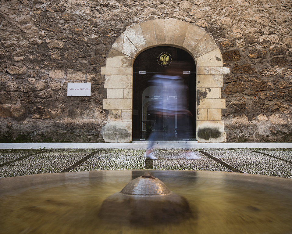 Patio del Hospital Real con fuente en el centro y puerta al fondo, a modo de ráfaga, una persona está pasando por la puerta