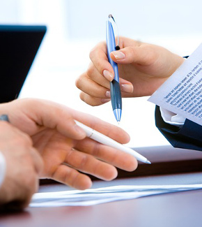 Hands holding pens and documents with a laptop in the background