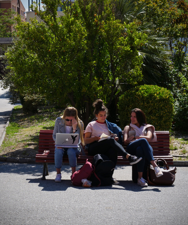 Tres estudiantes leyendo apuntes sentadas en un banco en el exterior de la Facultad de Ciencias del Deporte